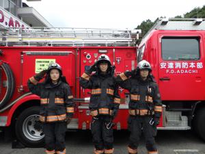 消防車の前で生徒３人が整列し防火服を着て敬礼